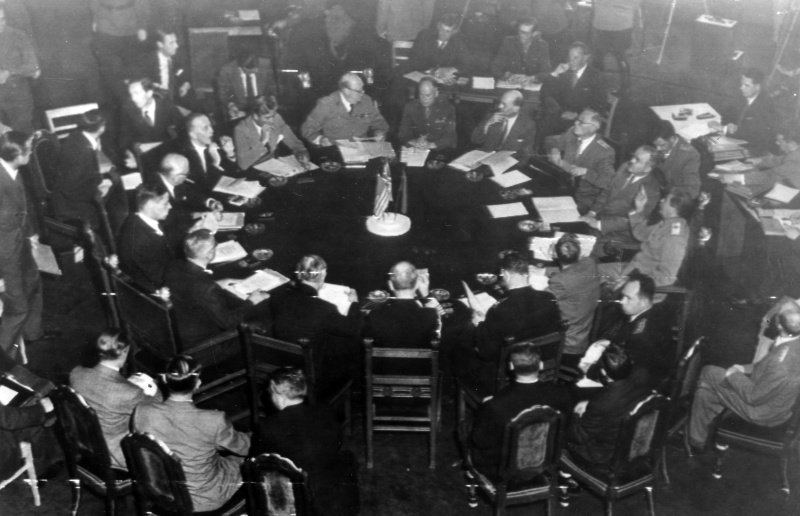 Black-and-white photograph of Allied political and military leaders seated around a large circular table during the 1945 Potsdam Conference. Delegates review documents, take notes, and engage in discussion inside a formal conference room. The round table emphasizes collective decision-making as representatives of the victorious powers negotiate postwar arrangements. The image captures the moment when wartime cooperation began giving way to uncertainty, laying the political and strategic foundations of the early Cold War.