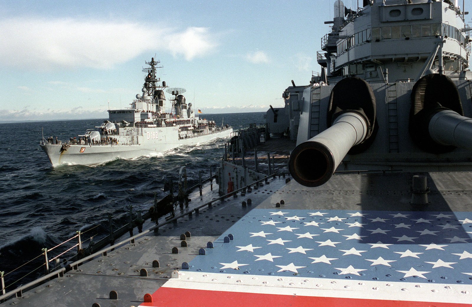 Color photograph of two Cold War–era warships operating together during a NATO exercise. In the foreground, the deck and gun turrets of the American battleship USS Iowa are visible. Nearby, the West German destroyer FGS Schleswig-Holstein sails alongside while refueling at sea during NATO Exercise Northern Wedding in 1986.